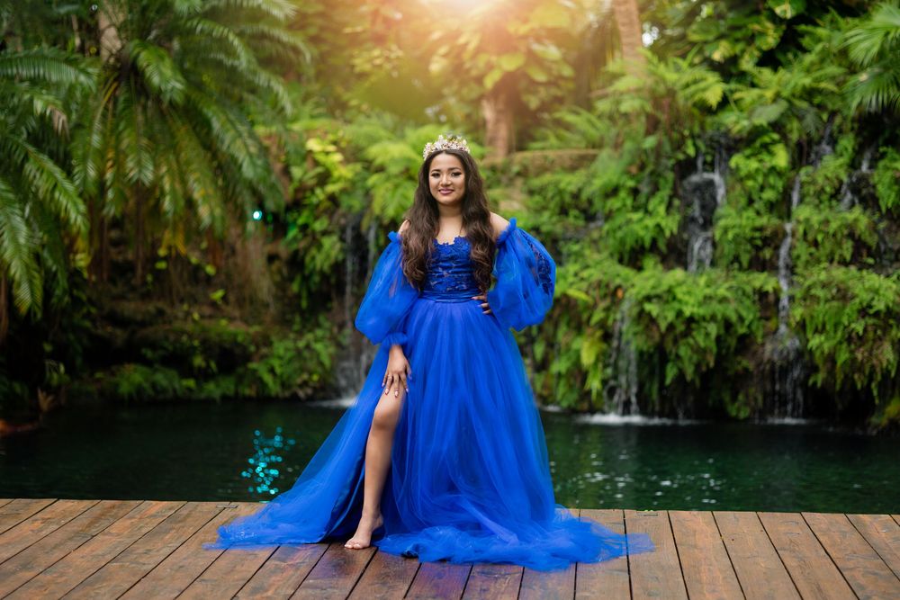 A quinceañera in a royal blue tulle gown with a crown stands on a deck near a tropical waterfall.