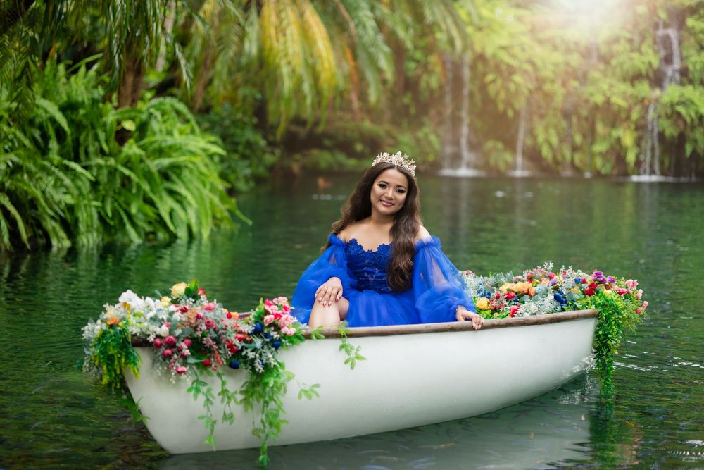 A quinceañera in blue gown sitting in a flower-adorned white boat on a serene green lake.