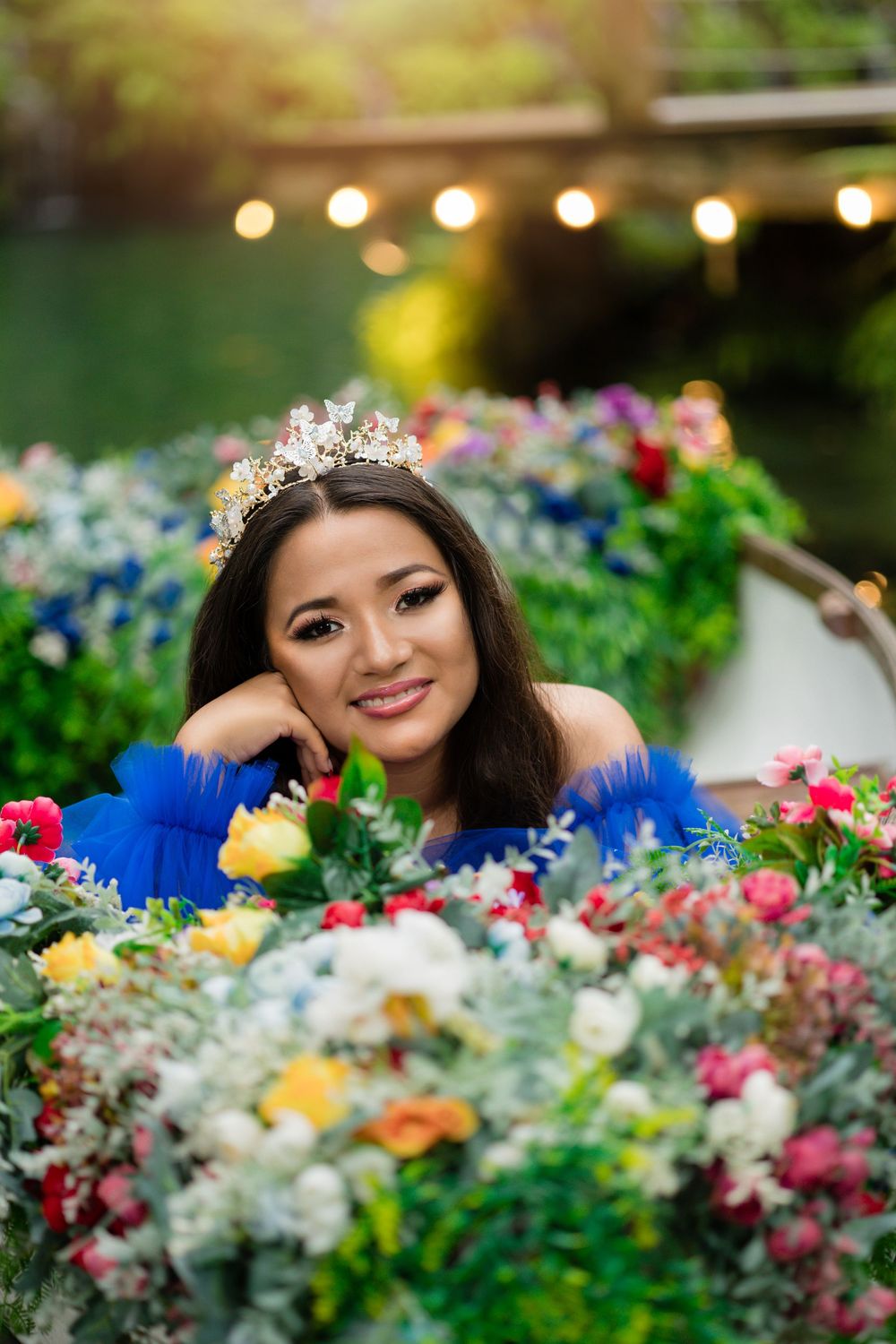 Quince Young woman in blue dress wearing a crown, posing among colorful flowers in a garden.