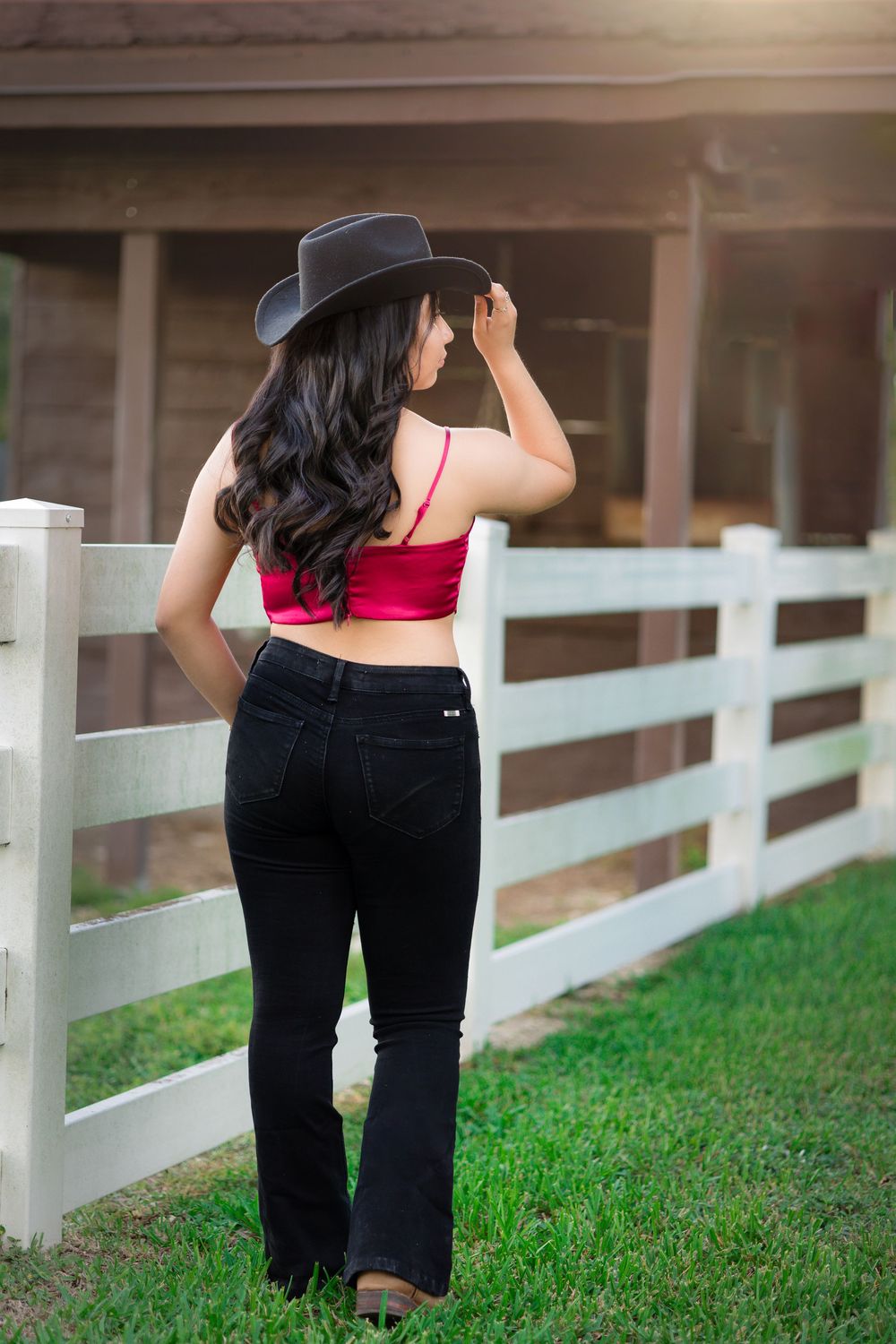 Young woman in pink crop top, black jeans, and cowboy hat stands by white fence at ranch.