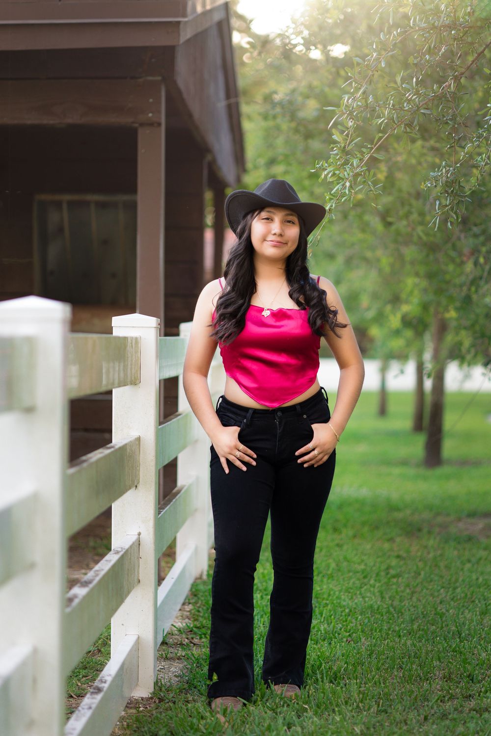 Smiling young woman in pink top and black jeans wearing cowboy hat poses by white fence.