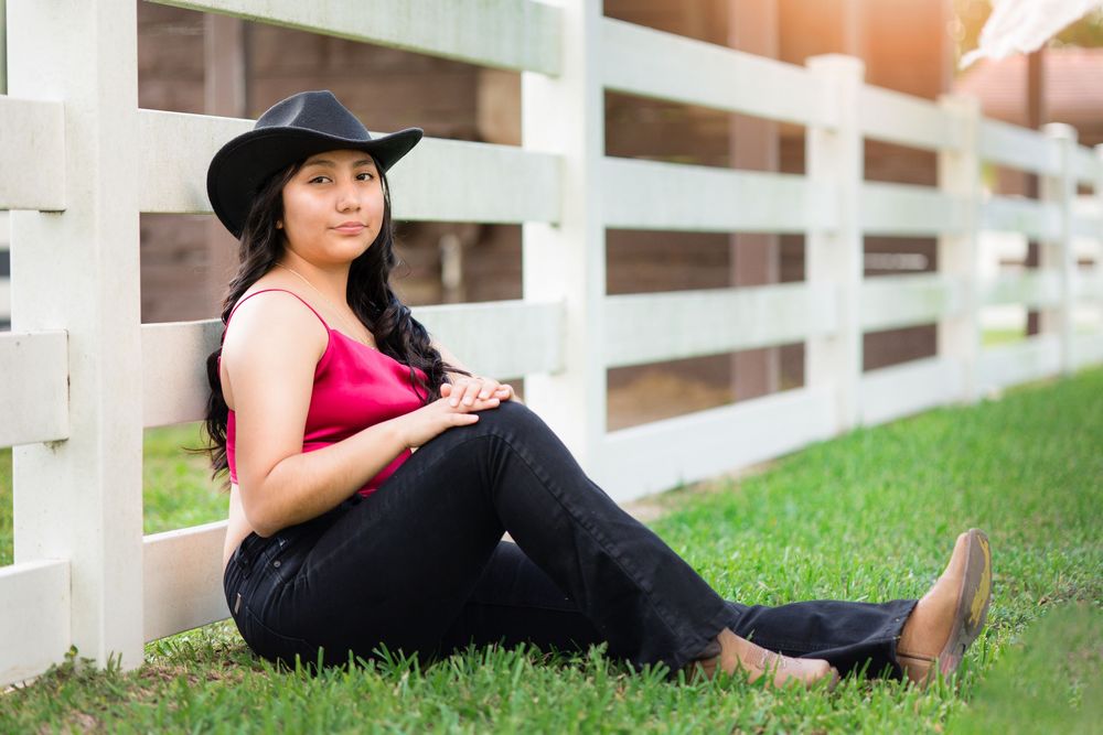 Young woman in pink top, black jeans, and cowboy hat sits relaxed against white fence on grass.