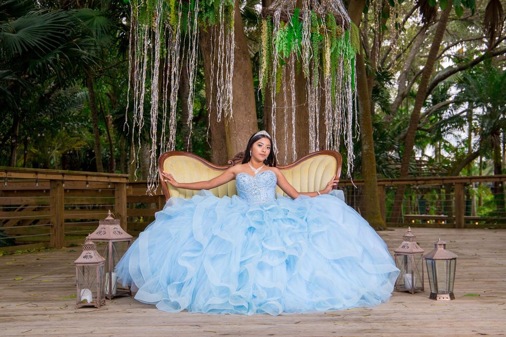 Young woman in a light blue quinceañera ball gown seated on a sofa on a tropical outdoor stage.