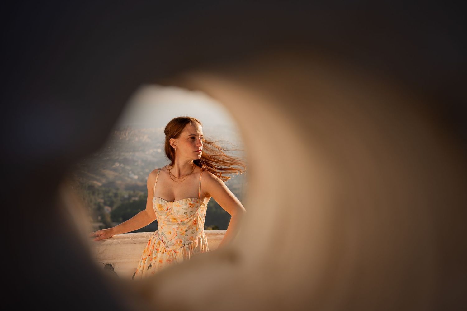 Through a circular window frame, a figure in a lace wedding dress poses against a blurred outdoor background in warm lighting.