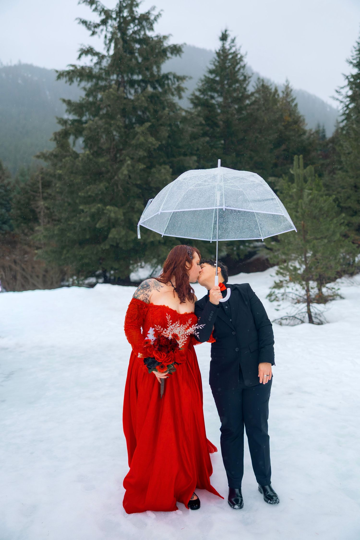 A romantic couple in black and red attire share an intimate moment by a misty lake with mountains in the background.