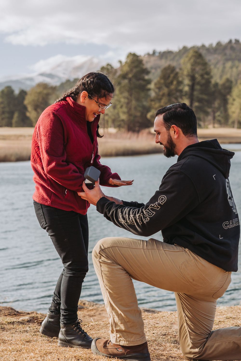 A sequence of proposal photos showing someone in red kneeling down to propose by a scenic lake on a sunny day.