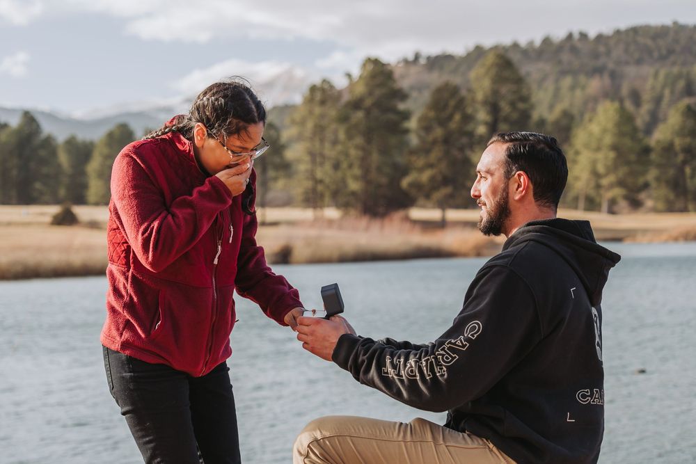 A proposal moment captured at a scenic lake with mountains and pine trees in the background.