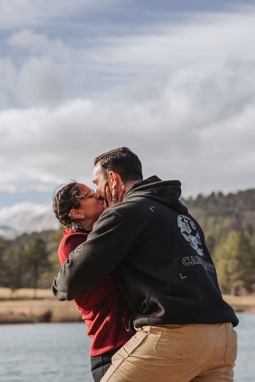 A couple in black and red clothing share an intimate moment while standing by a scenic lake with mountains in the background.