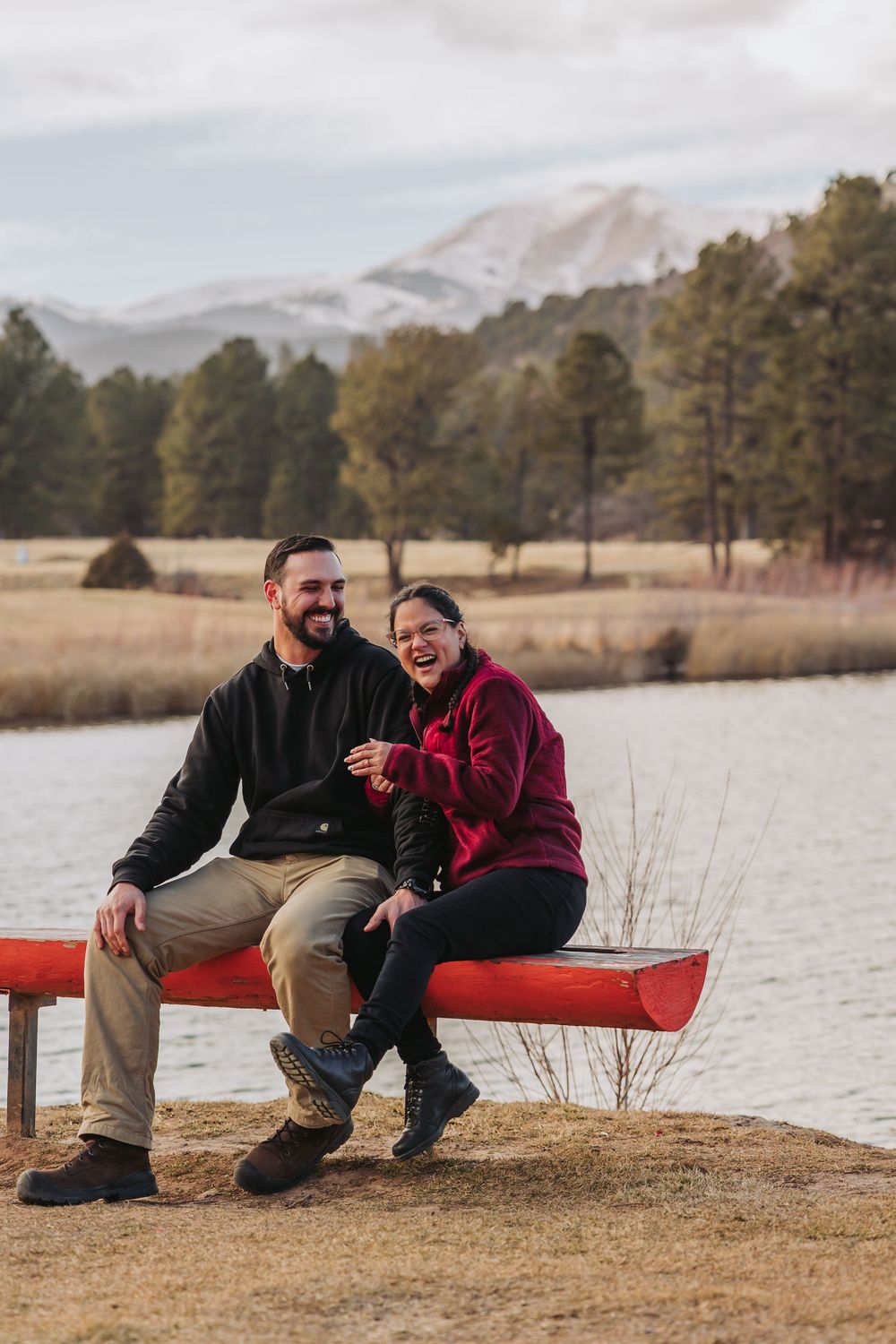 A couple sits together on a red bench near a lake with mountains and pine trees in the background.