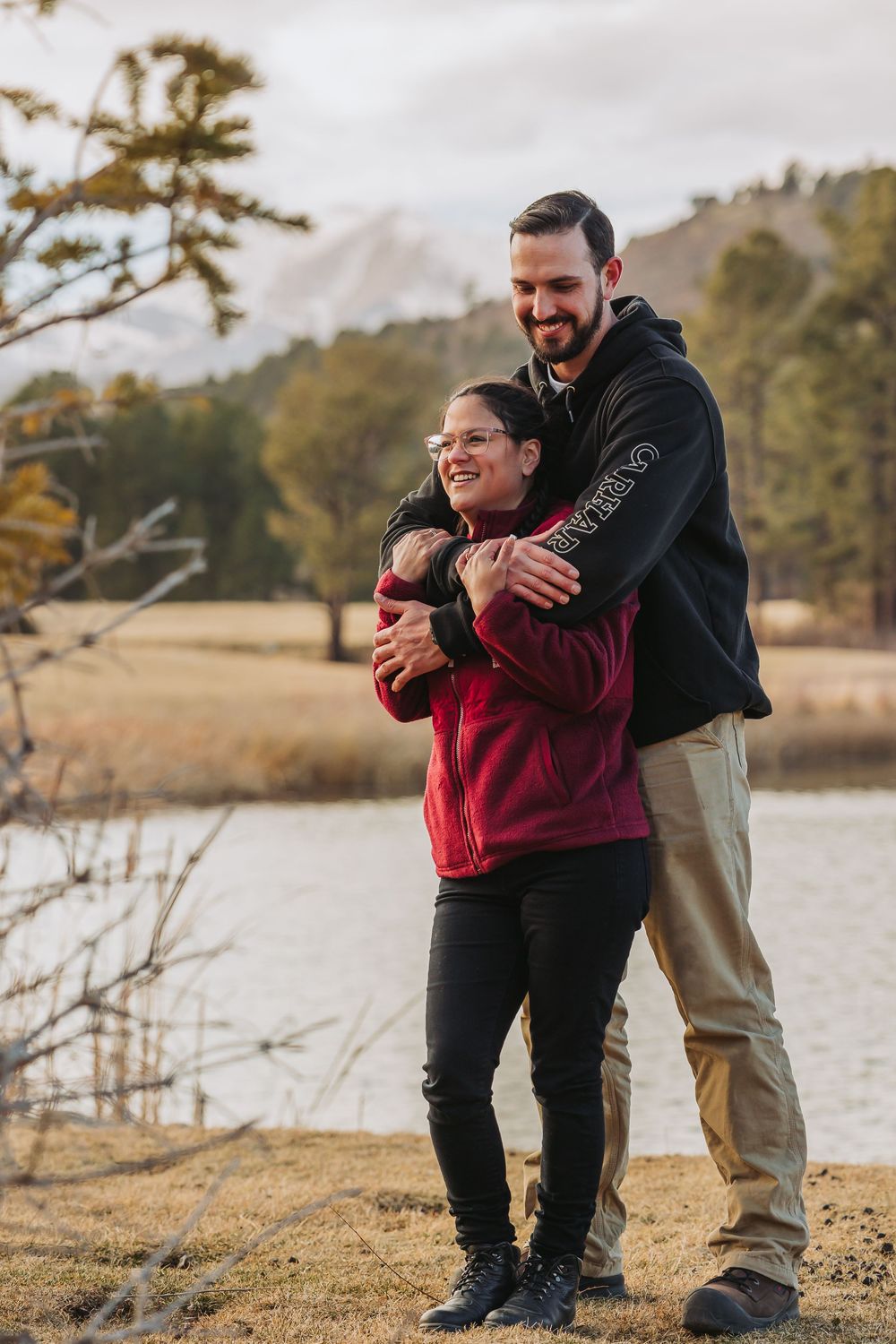 A heartwarming moment shared between two people embracing and laughing together by a scenic lake on an autumn day.