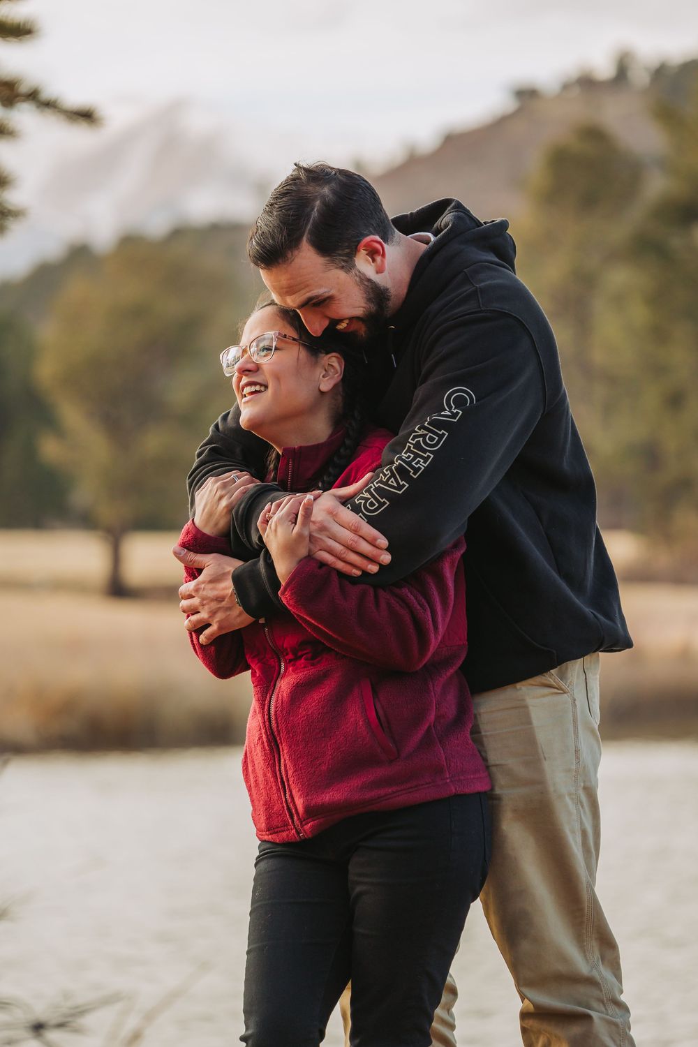 A couple embraces warmly by a serene lake during sunset, creating a tender romantic moment in nature.