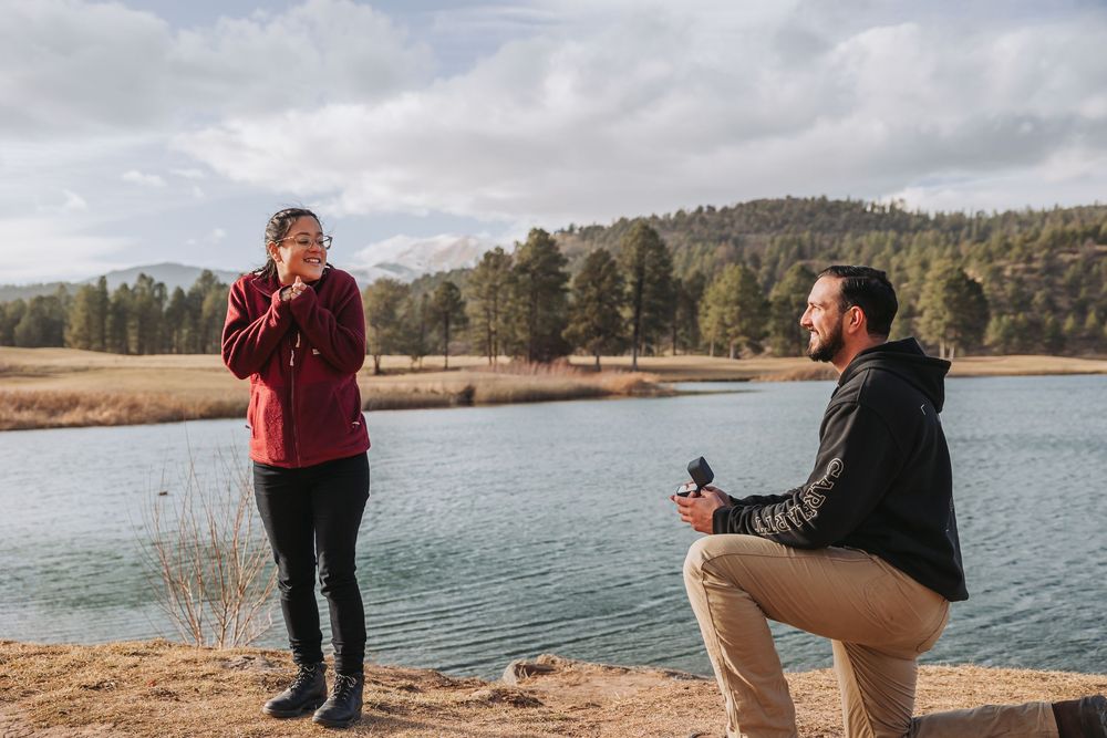 A romantic proposal moment unfolds on a scenic lakeside trail with mountains and water in the background.