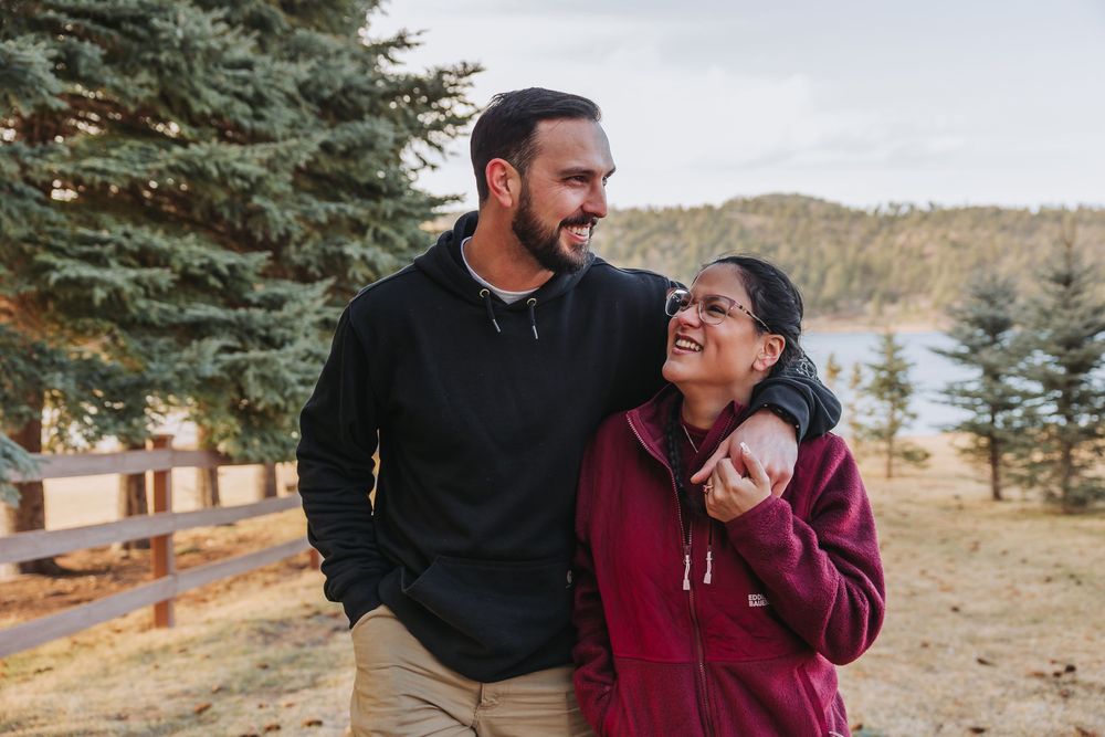 A couple shares a joyful moment together while standing near evergreen trees with a lake in the background.