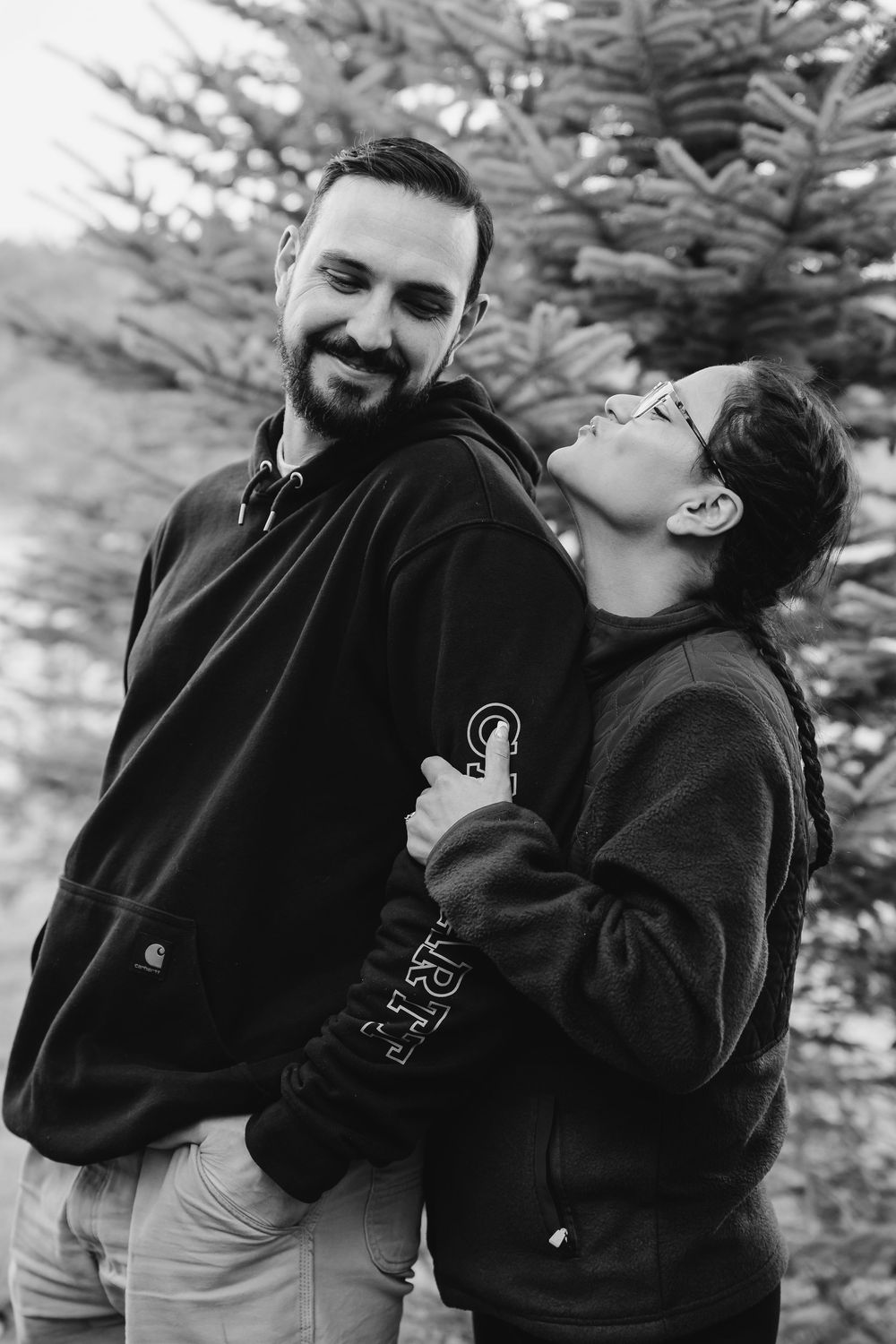 A playful couple in black hoodies share affectionate moments against an evergreen tree backdrop.