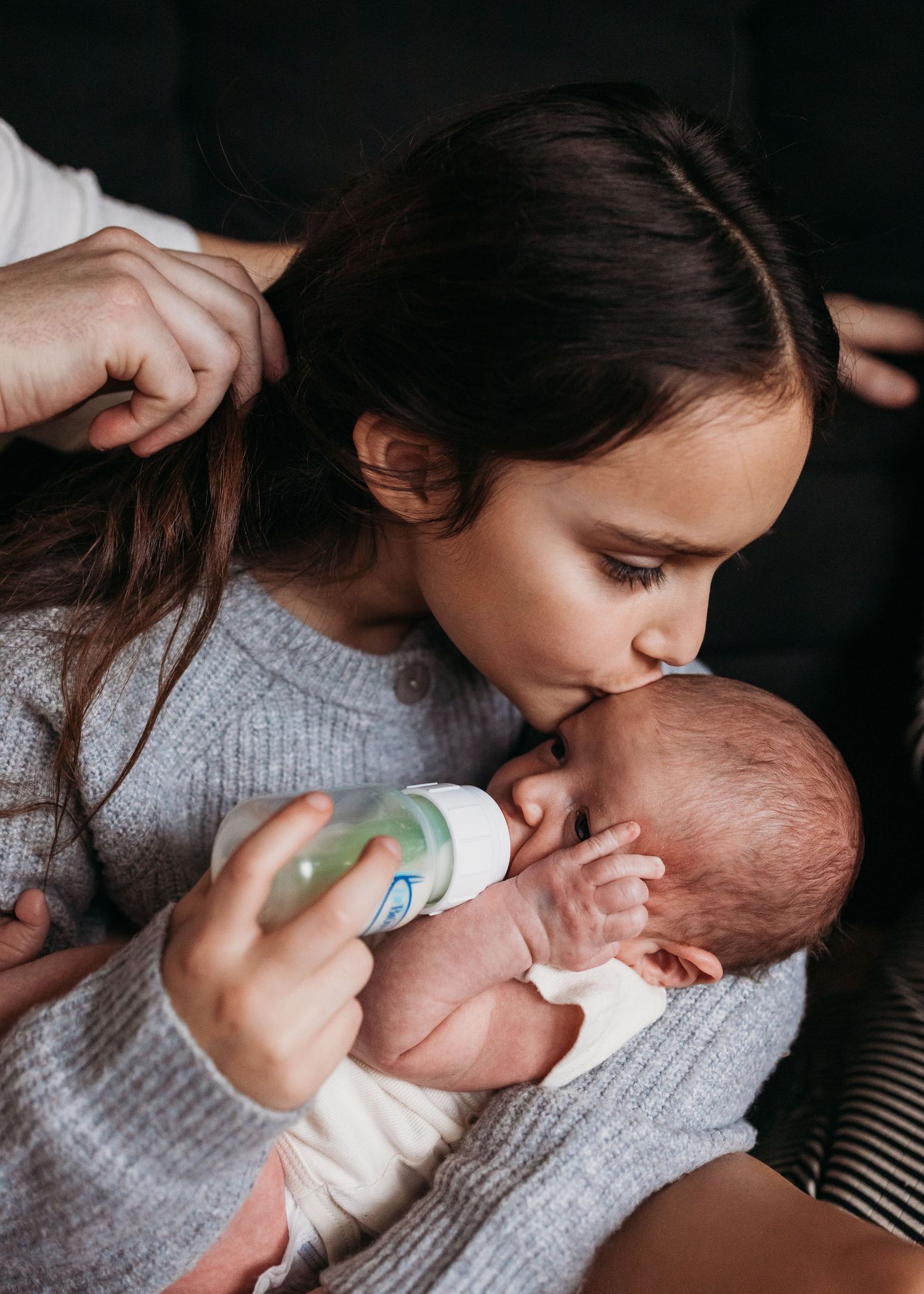 A child in a gray sweater tenderly kisses a baby while holding a bottle during feeding time.