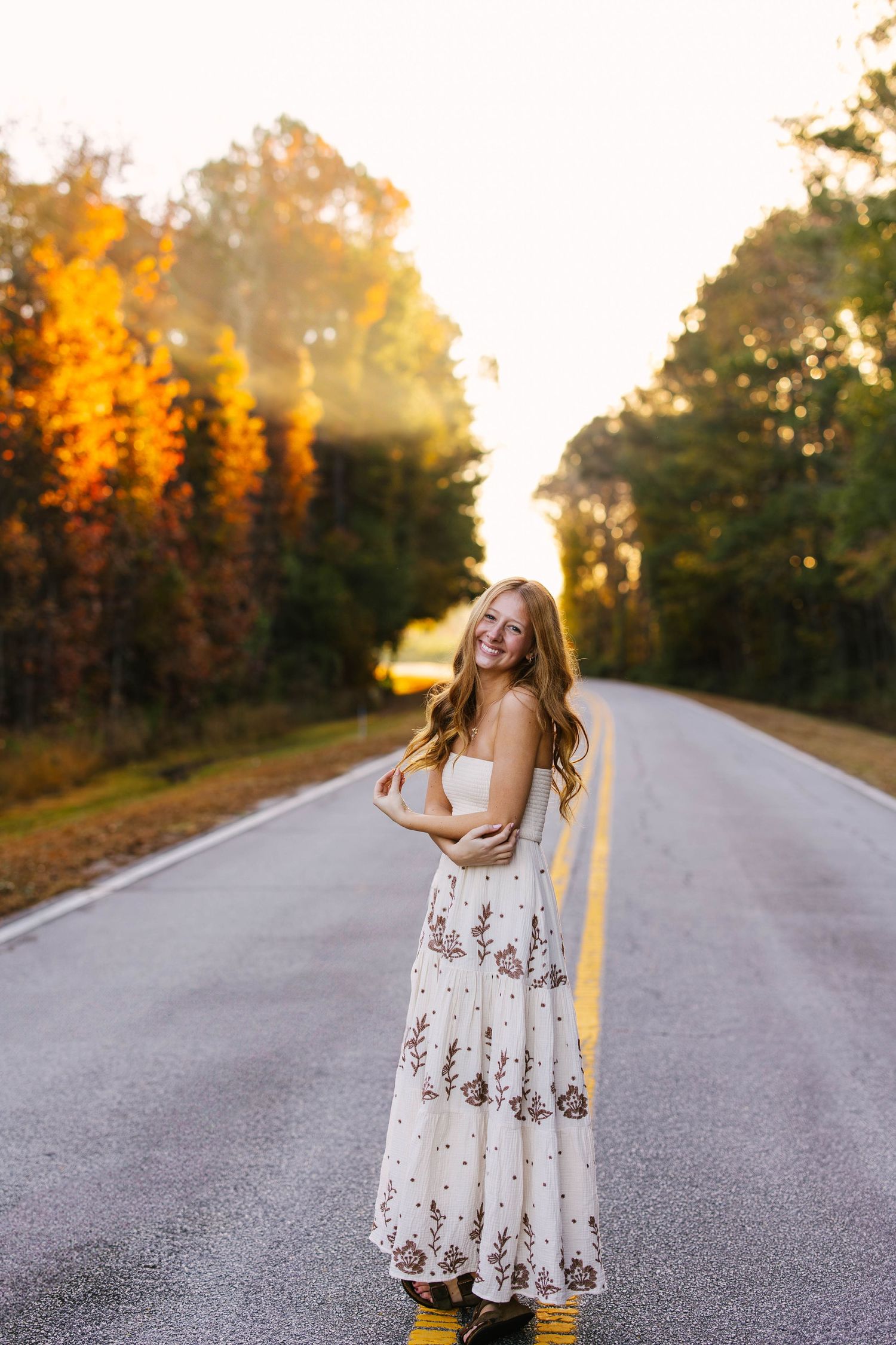 A figure in a white floral dress poses on an empty country road during golden hour with autumn trees in the background.