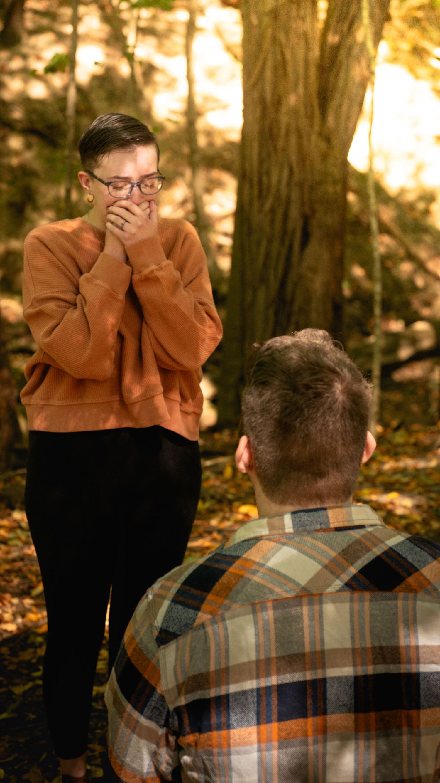A couple shares an emotional moment in a wooded area during autumn, with one person wearing a plaid shirt.