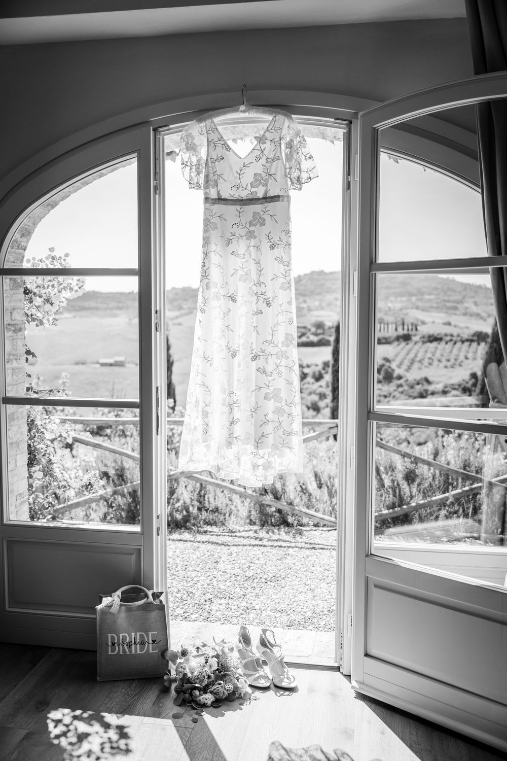 A delicate lace wedding dress hangs in an arched doorway with a scenic countryside view.