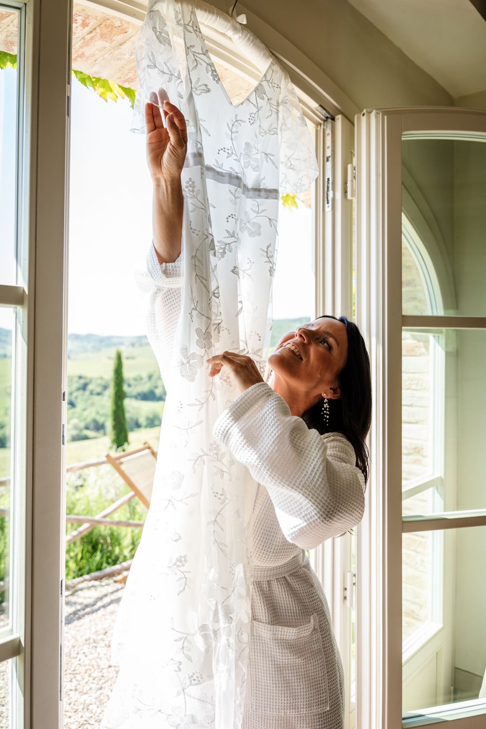 Someone in a white sweater hangs delicate lace curtains along a row of bright sunlit windows.
