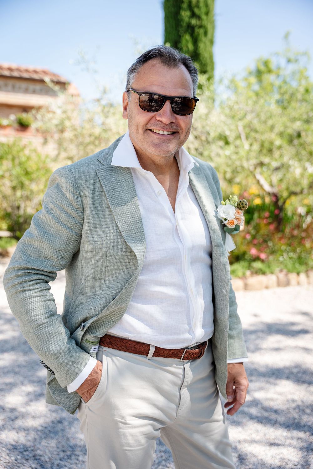 Stylish groom in light summer suit and sunglasses poses in Mediterranean garden setting.