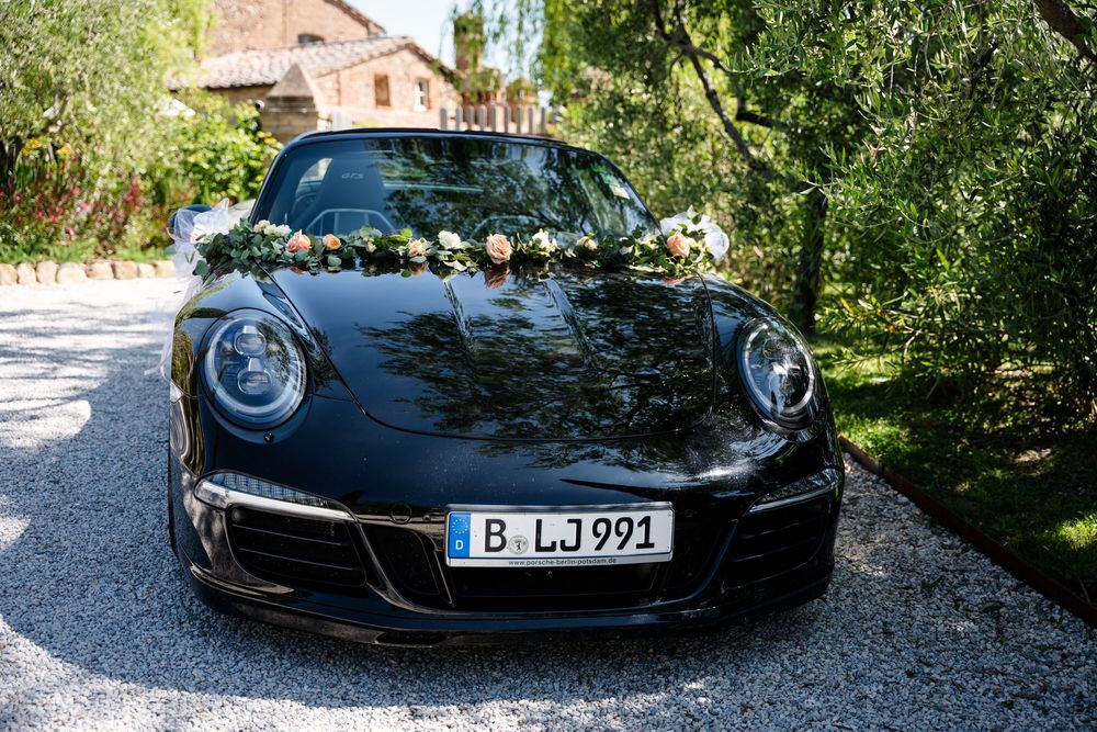 Black Porsche 911 decorated with white floral garland for a wedding celebration parked on a gravel driveway.