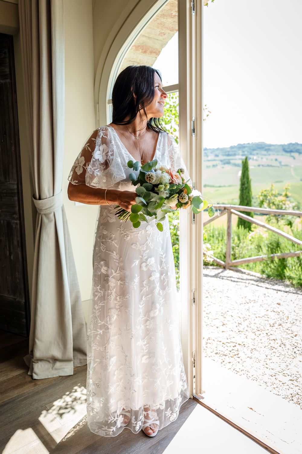 A bride in a lace wedding dress stands by an arched doorway holding a bouquet while gazing at a Tuscan countryside view.