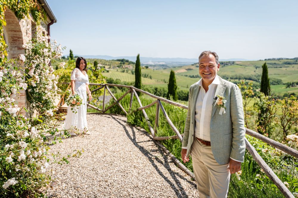 A wedding couple stands in a scenic Tuscan vineyard setting with rolling hills and cypress trees in the background.