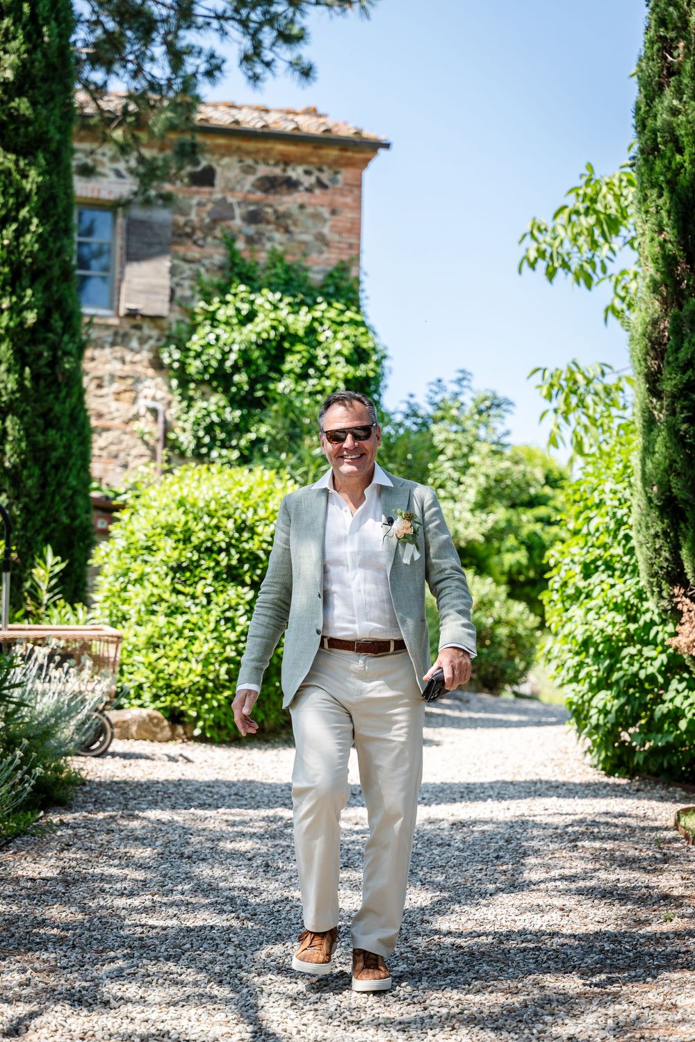 A groom in a light suit walks confidently down a cypress-lined gravel path.