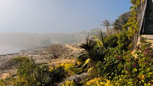 Panoramic view of a Mediterranean coastal landscape with mountains, palm trees, and white buildings against a blue sky.
