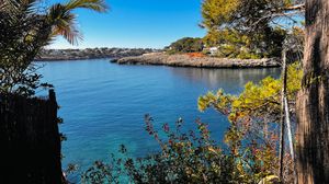 Wide panoramic shot of a Mediterranean marina with boats and coastal vegetation against blue waters.