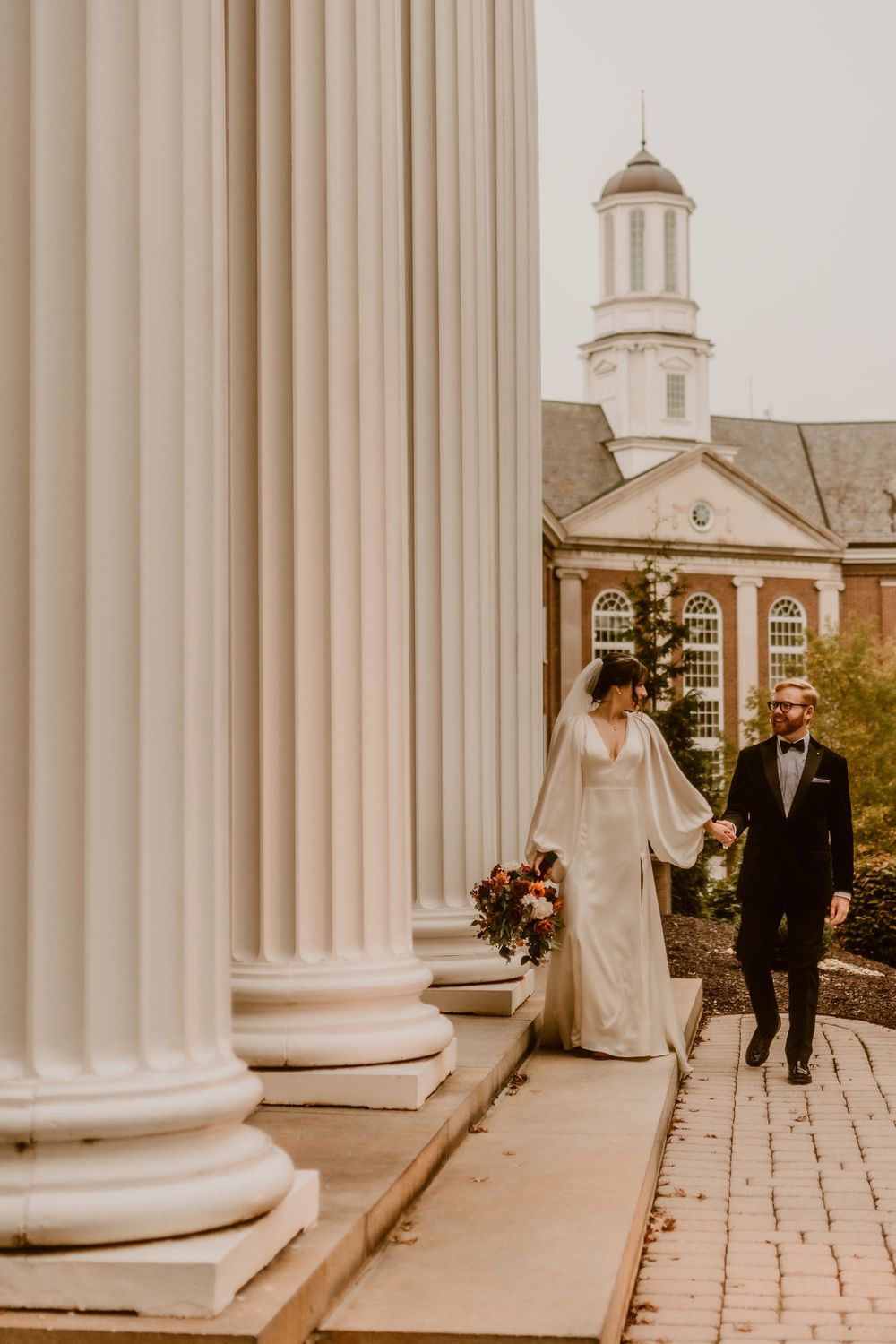 Couple walking between white columns with a domed building in the background.