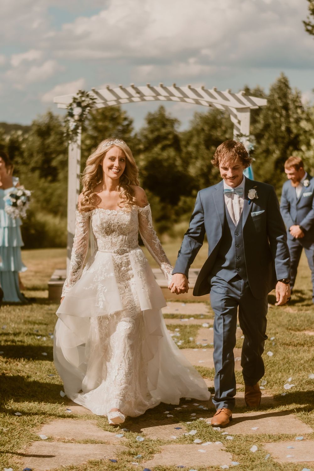 Wedding party walks along sunny garden path with bride in lace gown and bridesmaids in blue dresses.