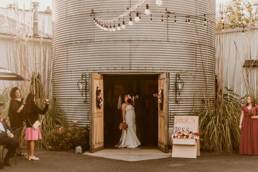 A wedding couple stands in the doorway of a rustic silo venue with string lights overhead.