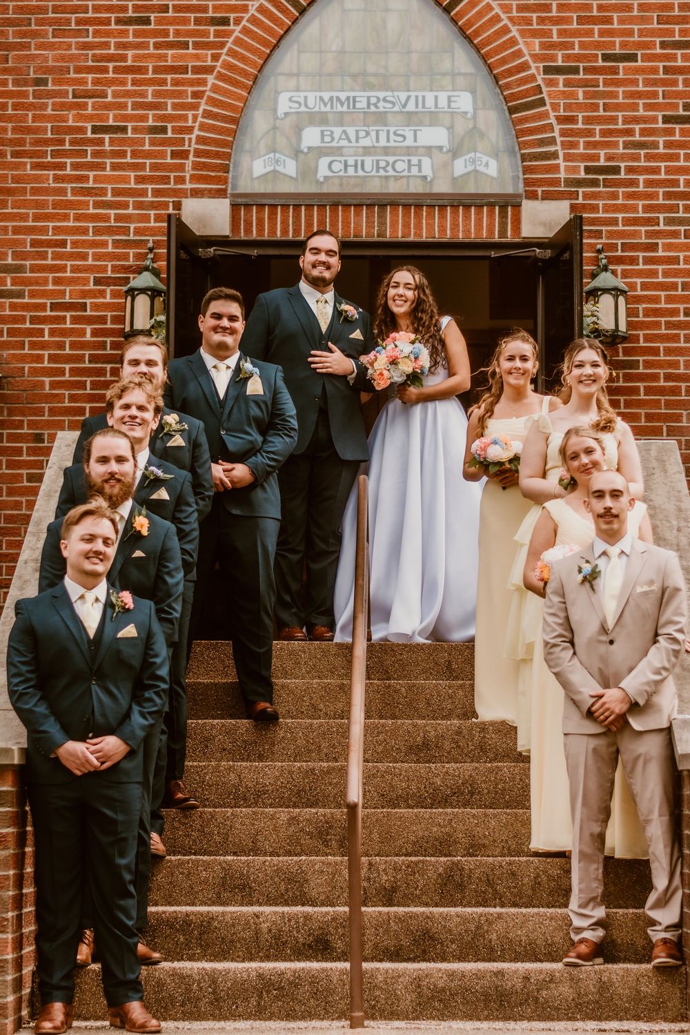Wedding party posing on church steps in front of brick facade and arched doorway.