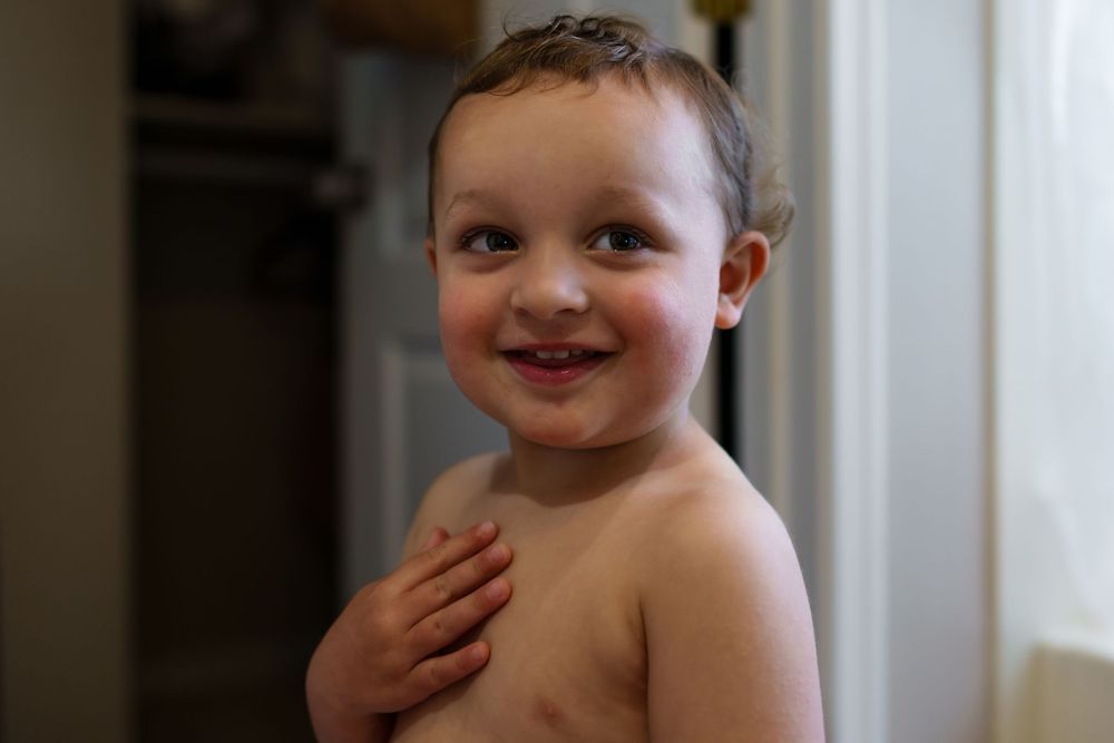 A smiling young child with bare shoulders stands indoors with a joyful expression.