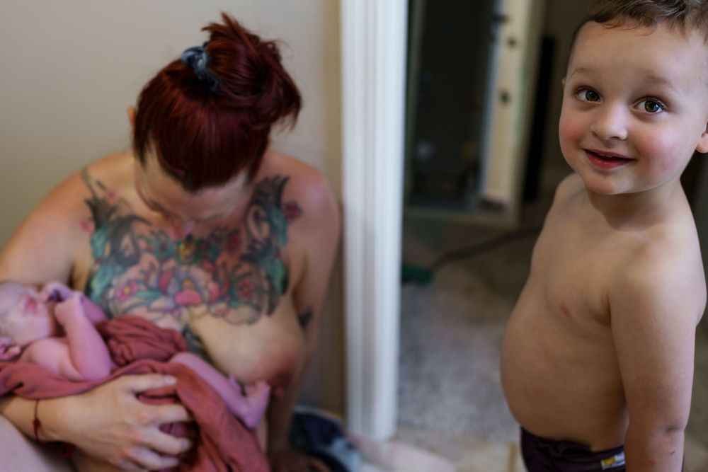A candid family moment shows a nursing parent with a baby while a smiling toddler stands nearby.
