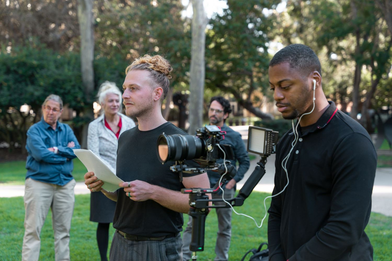 Film crew members operate a professional camera while shooting outdoors in a park setting.