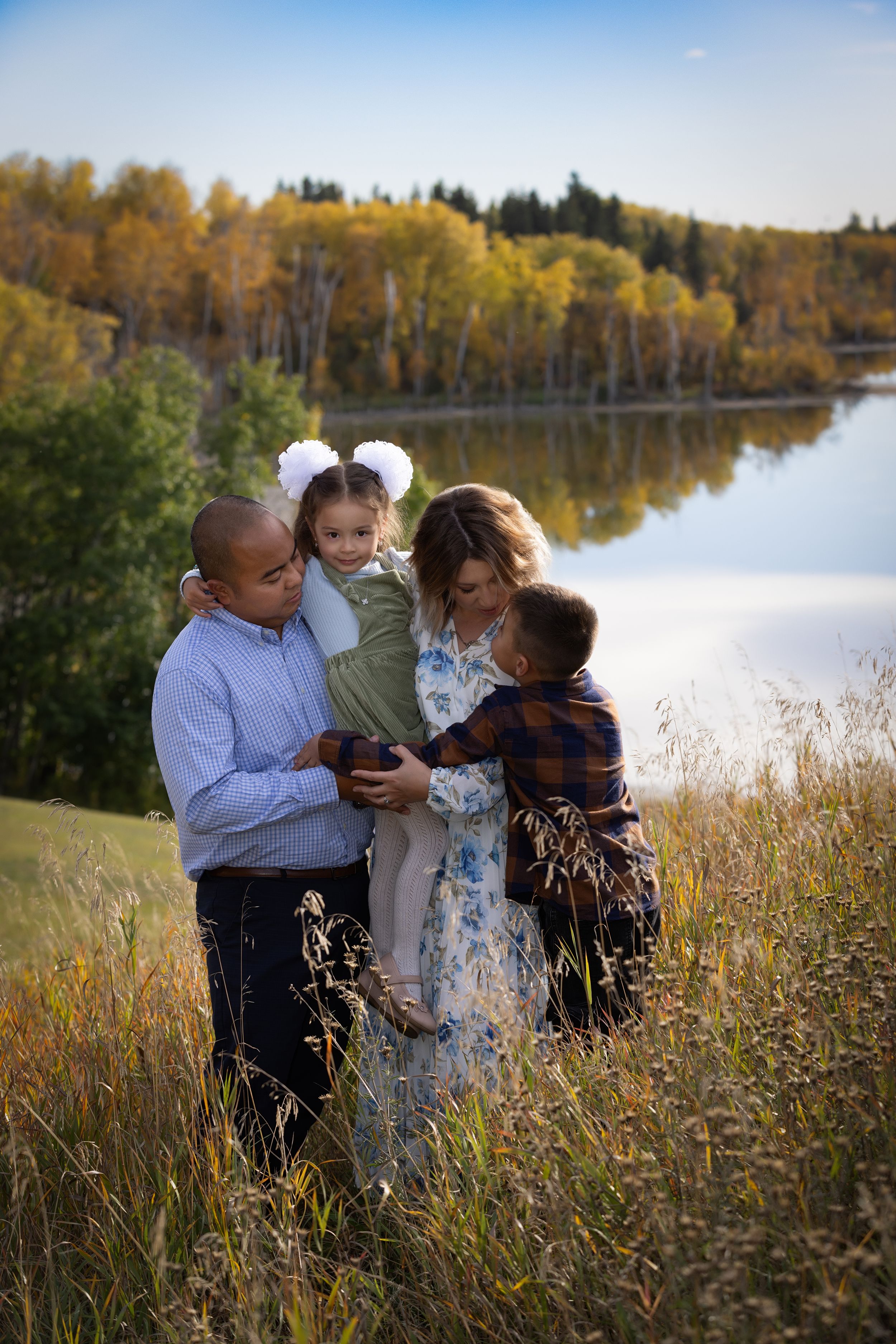 Family shares tender moments together in tall autumn grass by a serene lake during golden hour.