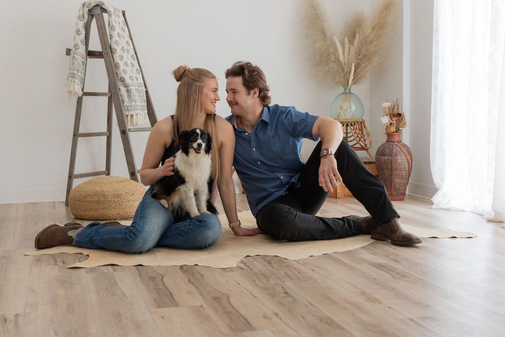 A couple sits on hardwood floor with their dog while holding wine glasses in a bright minimalist room with a ladder.