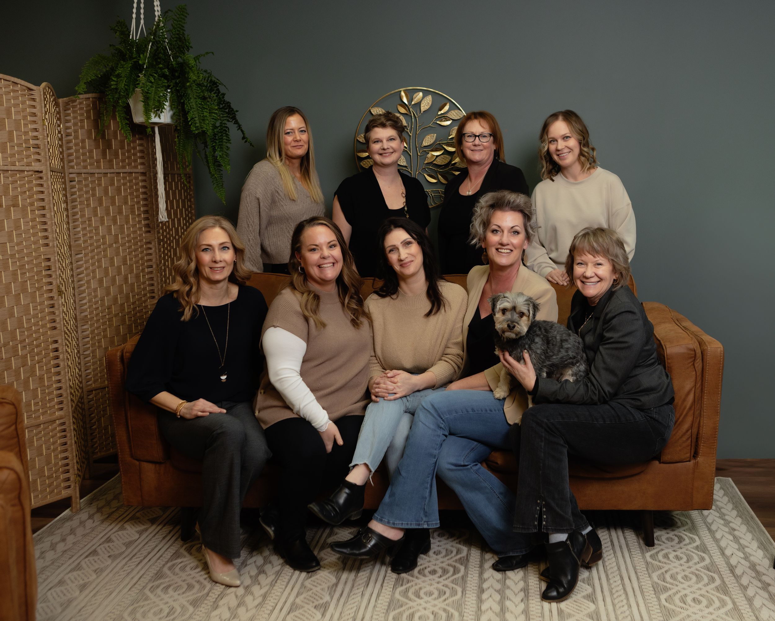 A group of people sitting together on a brown couch in a cozy studio setting with woven wall decor and plants.