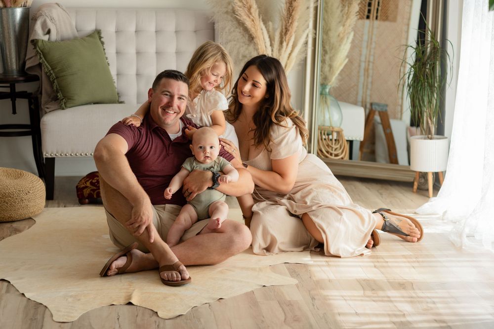 A family sits together on a cream-colored rug in a cozy living room with natural decor and sunlight.