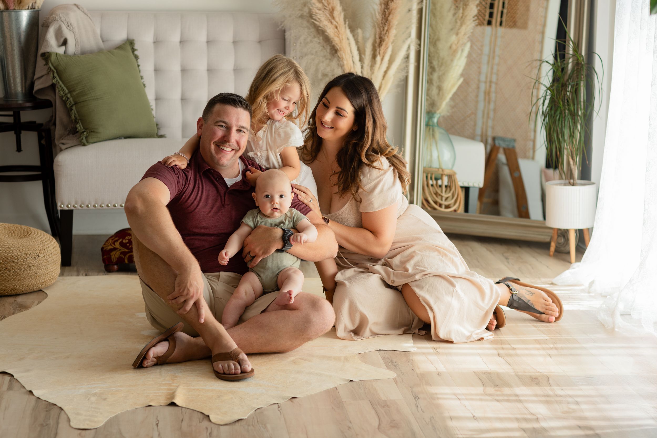 A family sits together on a cream-colored rug in a cozy living room with natural decor and sunlight.