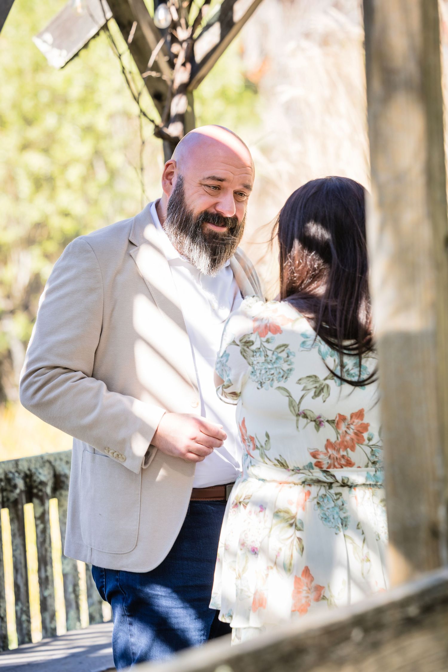 A couple shares a romantic moment outdoors in a wooded area with sunlight filtering through the trees.