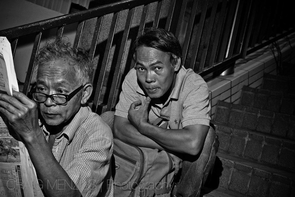 Two older Asian men sitting on stairs, one reading a newspaper, in black and white street photography.