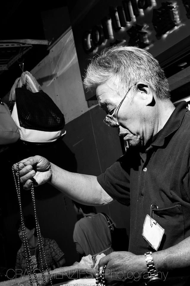 Black and white photo of an older man examining beaded jewelry in a shop.