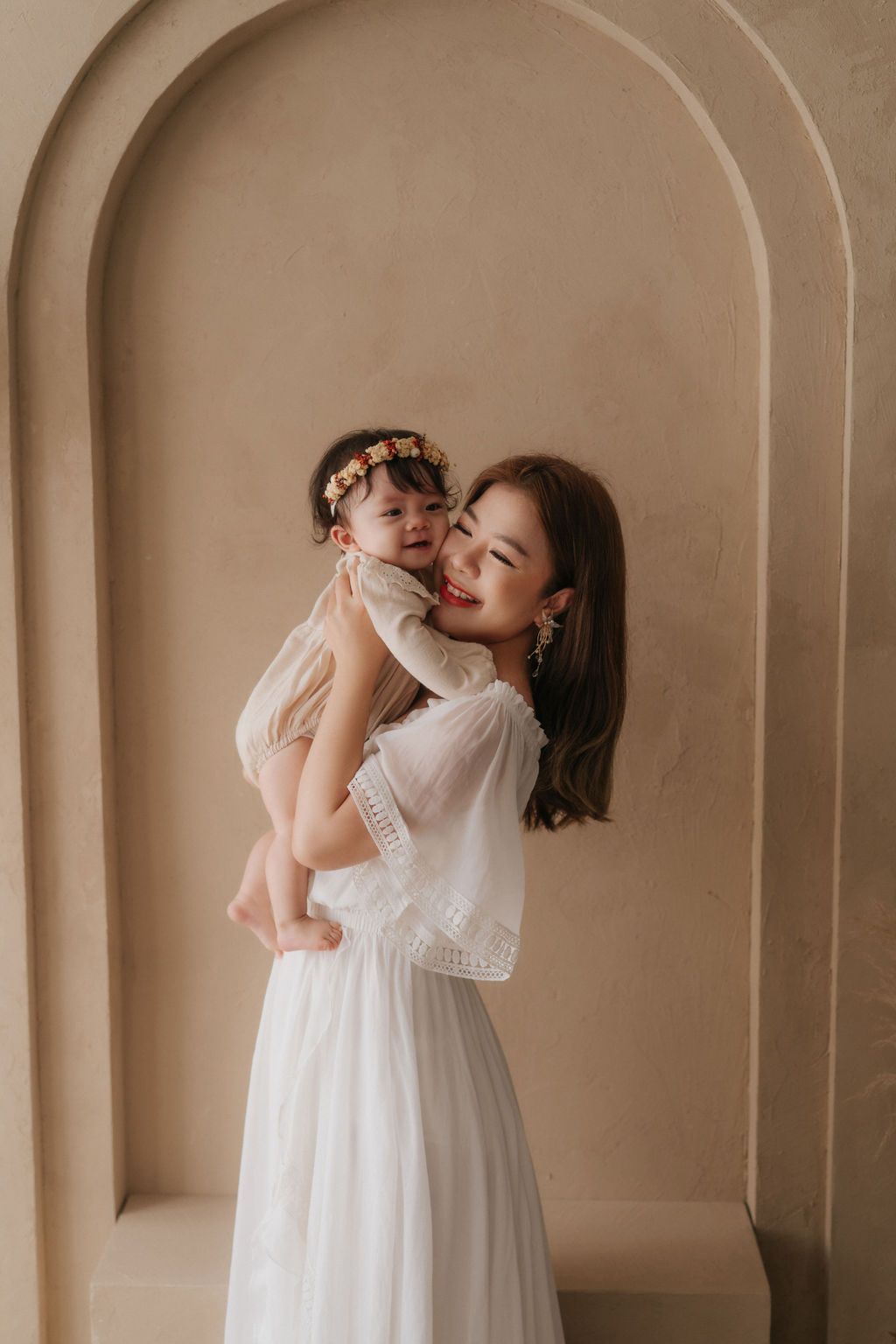 Woman in white lace dress holds smiling baby with floral crown against beige arched wall backdrop.身穿白色蕾絲洋裝的女子抱著戴著花冠的微笑嬰兒，背景是米色拱形牆。