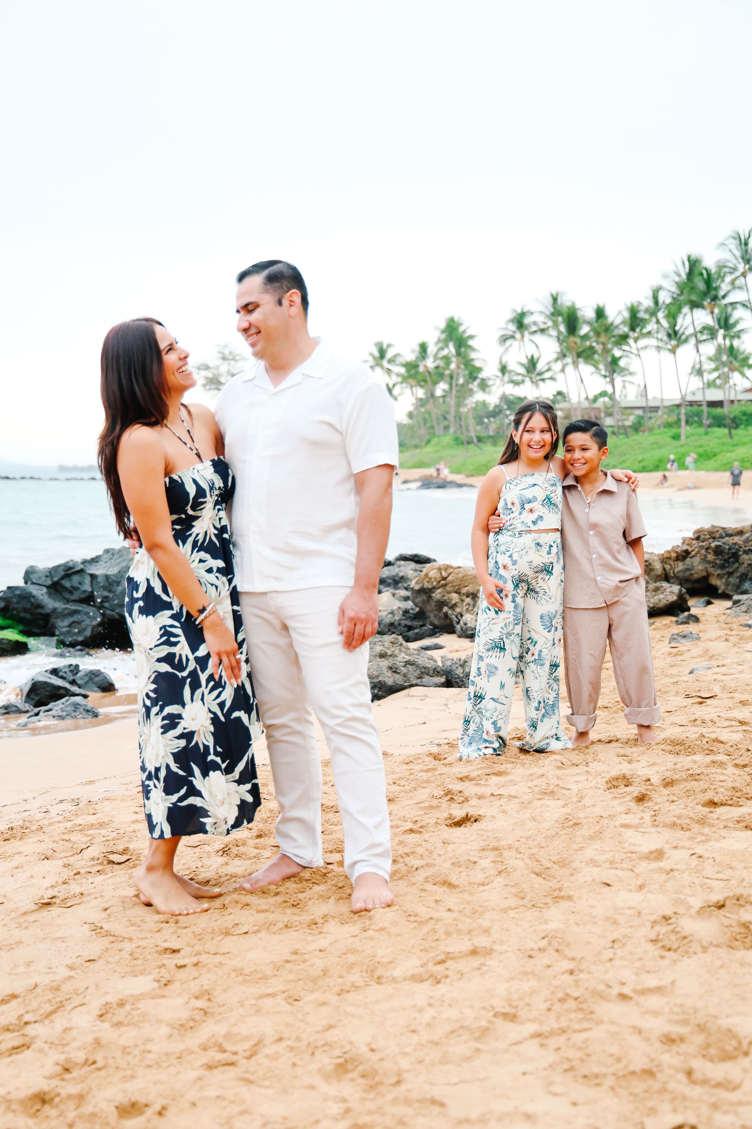 A couple shares romantic moments walking along a sandy beach with rocks and palm trees in the background.