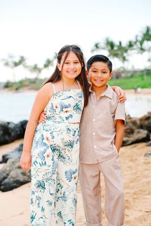 A couple in coordinating outfits of floral dress and beige suit pose together on a tropical beach with palm trees.