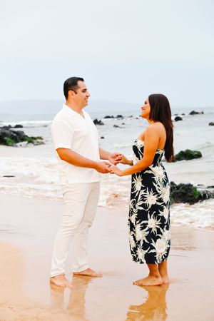A couple in matching white and floral outfits share an intimate moment on a beach with black rocks.