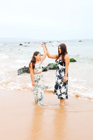 Two people in floral dresses hold hands and dance together at the ocean's edge.